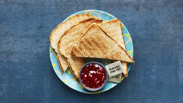 Toasted bread slices on a patterned plate with a side of red jam and butter packets on a blue textured background.