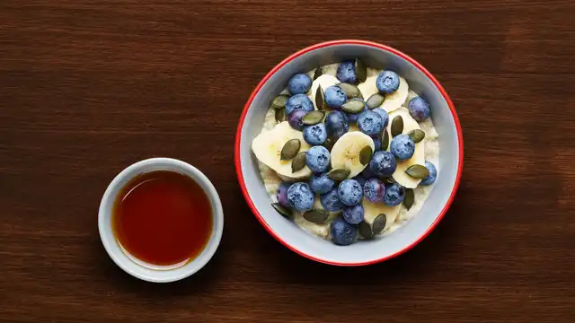 Bowl of porridge topped with blueberries, banana slices, and pumpkin seeds, next to a small bowl of maple syrup on a wooden table.