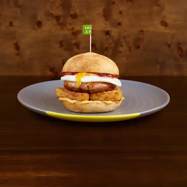 Burger with chicken, egg, hash brown on a bun, garnished with a green "Lemon & Herb" flag, served on a grey plate.