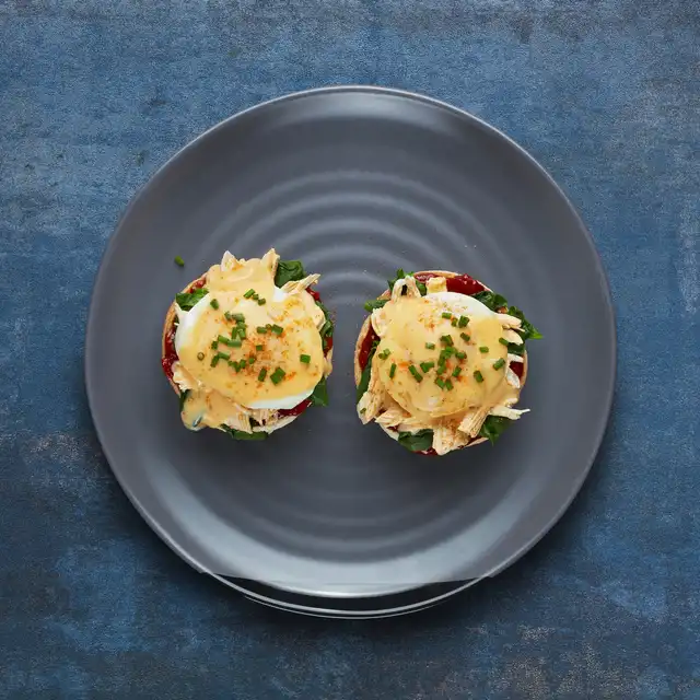 Two eggs Benedict on a dark plate, topped with creamy hollandaise, shredded chicken, and a sprinkle of chives.