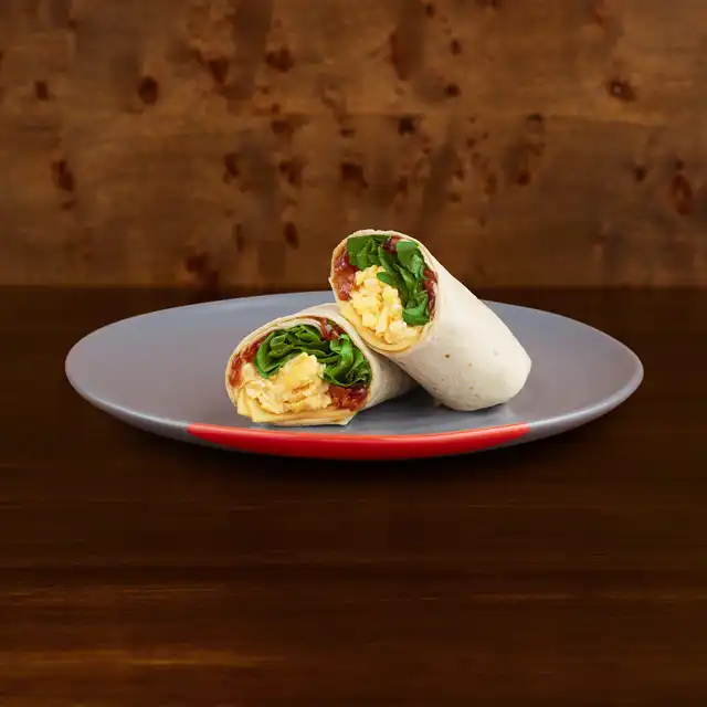 A breakfast wrap with scrambled eggs, cheese, lettuce, and tomato is sliced in half and served on a grey plate with a red rim.