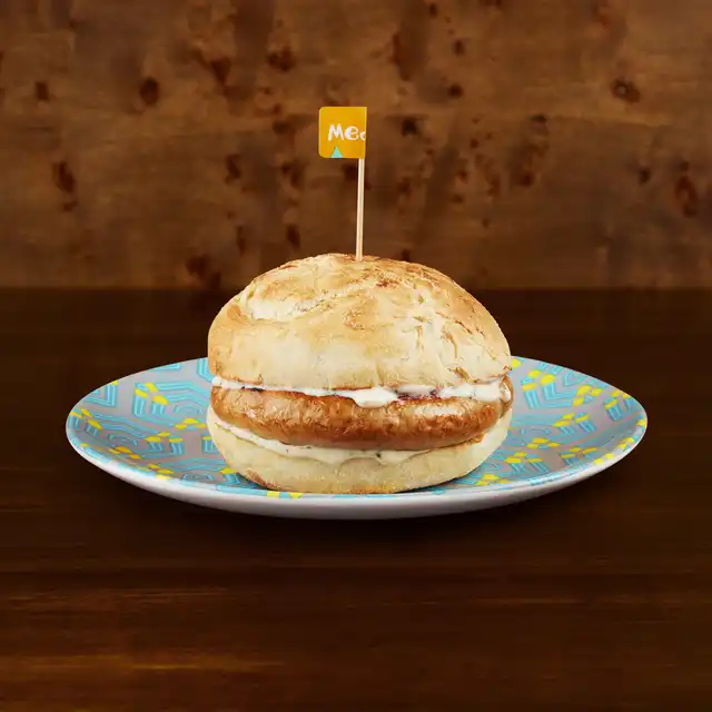 A grilled sausage burger with sauce on a colourful plate, marked with a small "Mega" flag, set against a wooden background.